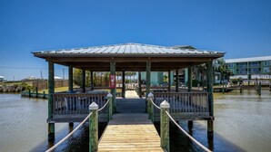 En träbrygga som leder fram till en överbyggd pergola med räcken, omgiven av stilla vatten och byggnader i bakgrunden.