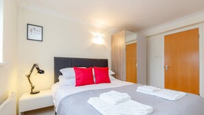 Ein ordentlich gemachtes Bett mit weißen und roten Kissen, einer Nachttischlampe und gefalteten Handtüchern auf einem weißen Nachttisch.