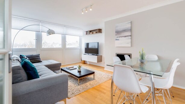 Ein modernes Wohnzimmer mit grauem Sofa, einem Glastisch, weißen Stühlen, einem Flachbildfernseher und einem großen Fenster.