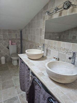Un baño con dos lavamanos grandes y redondos, de color beige, un espejo encima y un inodoro con un revestimiento de azulejos.