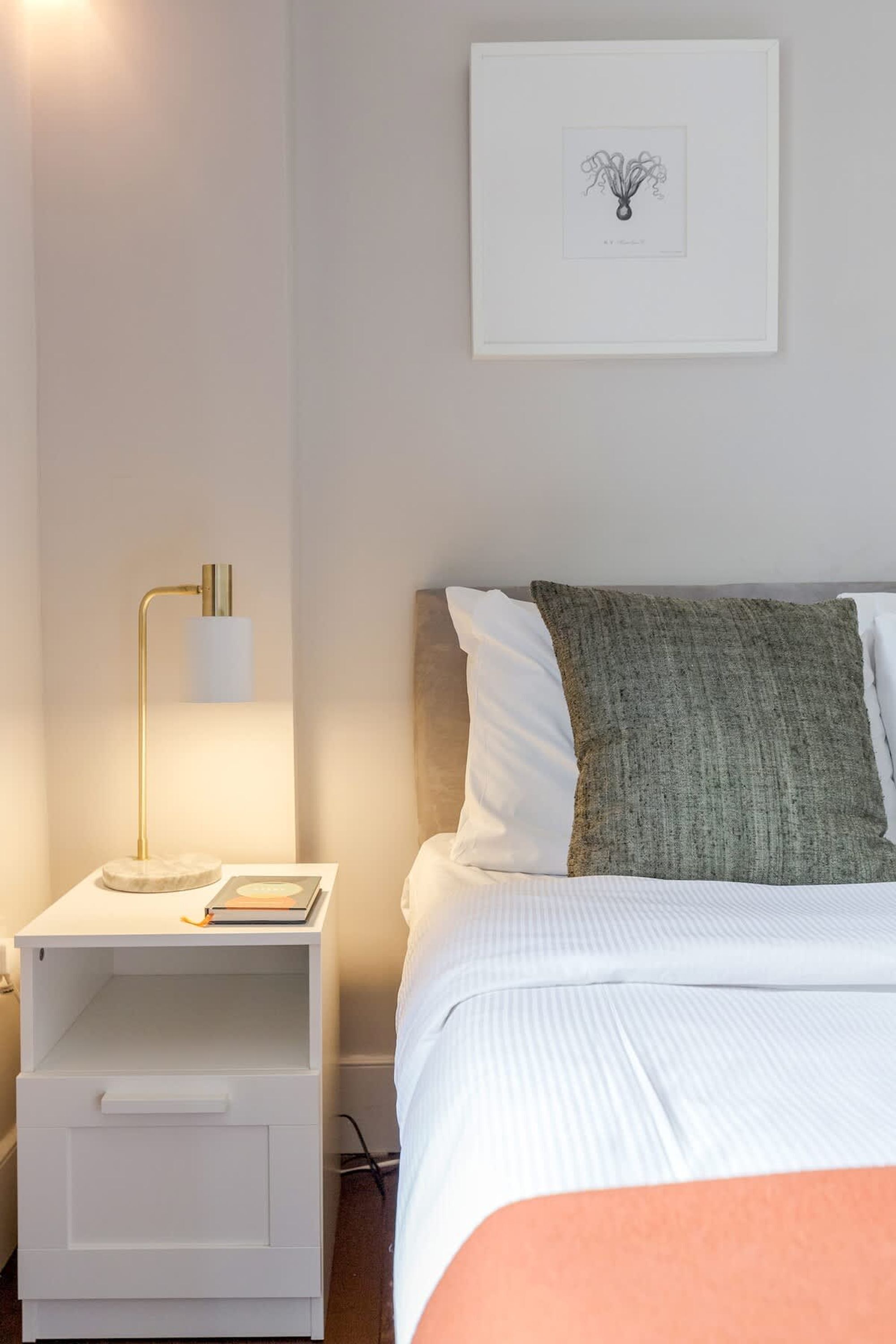 Een netjes opgemaakt slaapkamer met een bed, een nachtkastje met een lamp en een lijstje aan de muur.