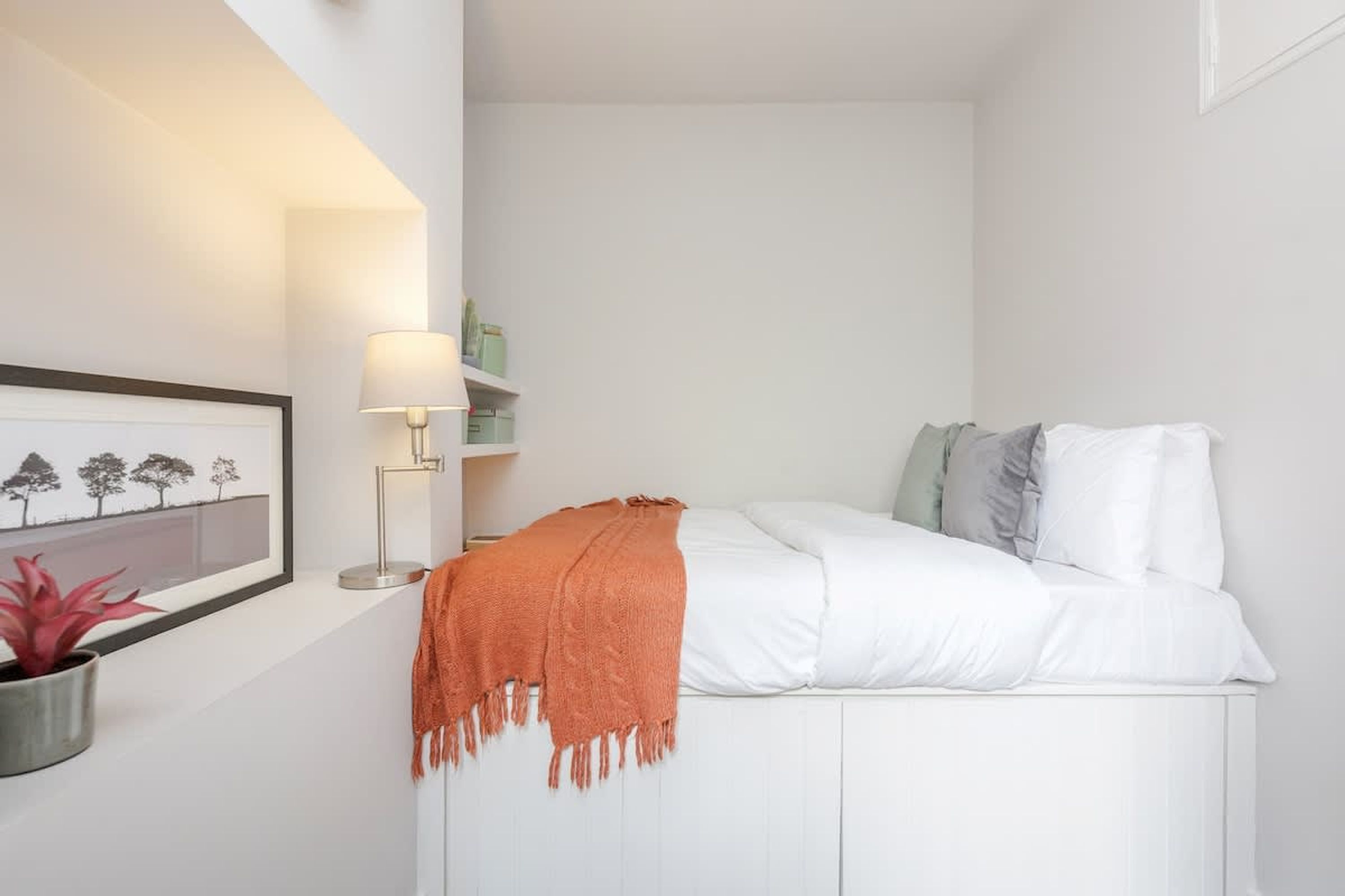 Un dormitorio ordenado con una cama, una lámpara de noche, un cuadro enmarcado y una planta en maceta.
