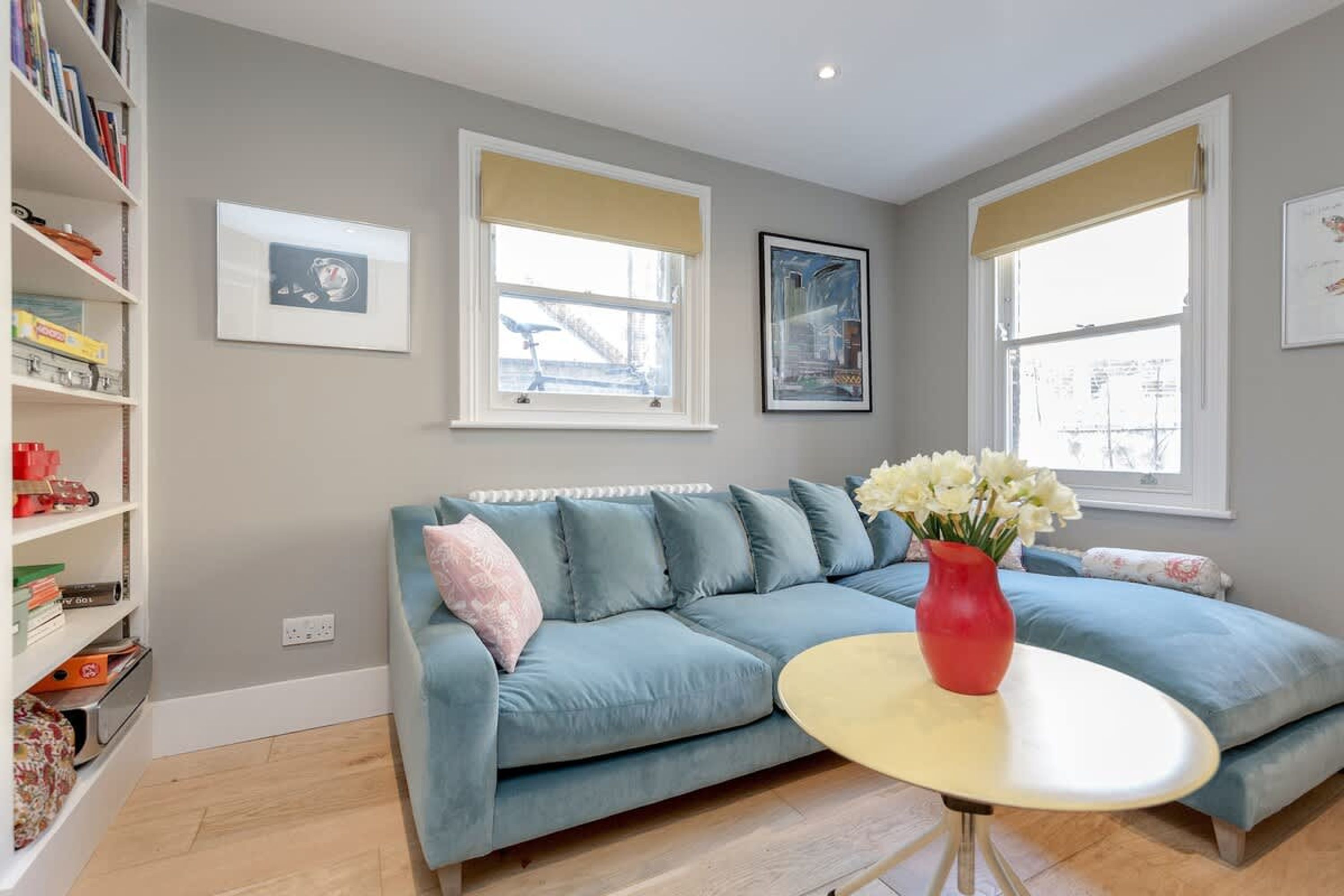 A living room with a blue sofa, a red vase with flowers, and a white round table. There are two windows with beige blinds, a bookshelf, and framed pictures on the wall.