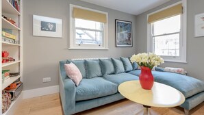 A living room with a blue sofa, a red vase with flowers, and a white round table. There are two windows with beige blinds, a bookshelf, and framed pictures on the wall. - 3bed House for an Authentic stay near river metro (Greater London)