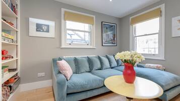 A living room with a blue sofa, a red vase with flowers, and a white round table. There are two windows with beige blinds, a bookshelf, and framed pictures on the wall.