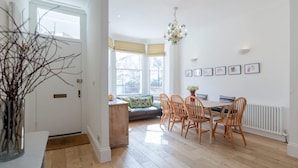 A bright dining area with wooden flooring, a table with chairs, a chandelier, and a sofa with cushions. - 3bed House for an Authentic stay near river metro (Greater London)