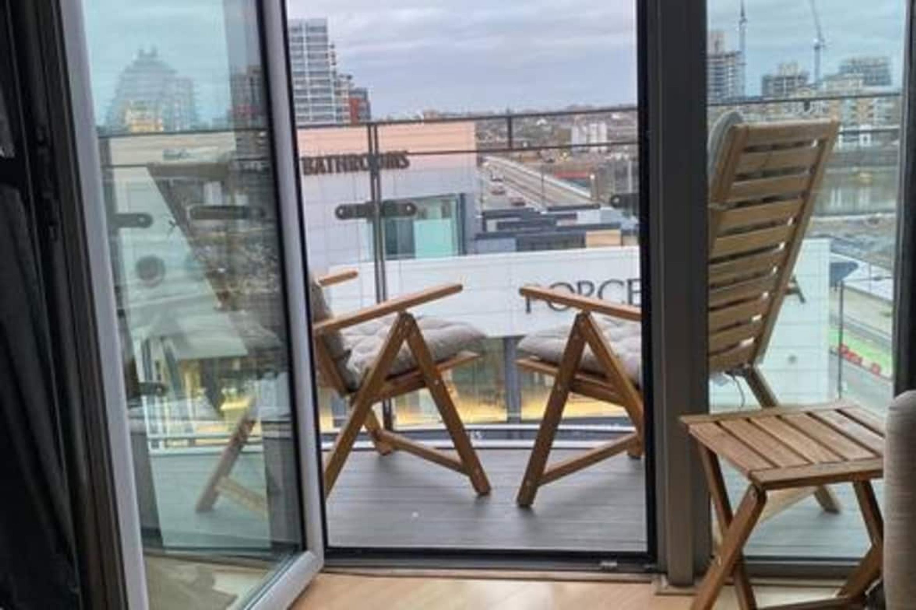 Un balcon avec deux chaises en bois et une petite table, donnant sur un paysage urbain avec des immeubles et un pont.