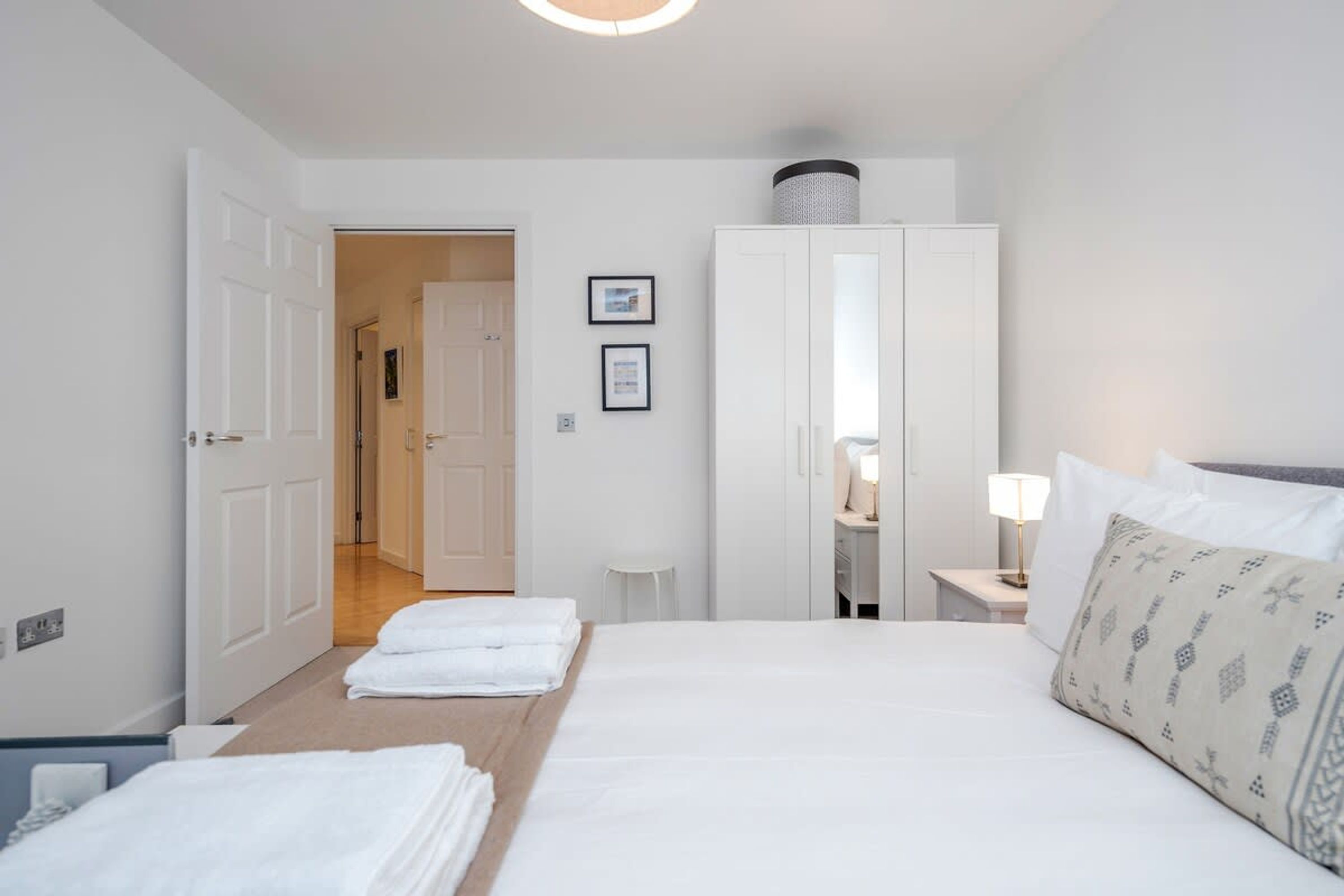 A hotel room with a bed, a wardrobe, a desk with a lamp, and a door leading to another room.