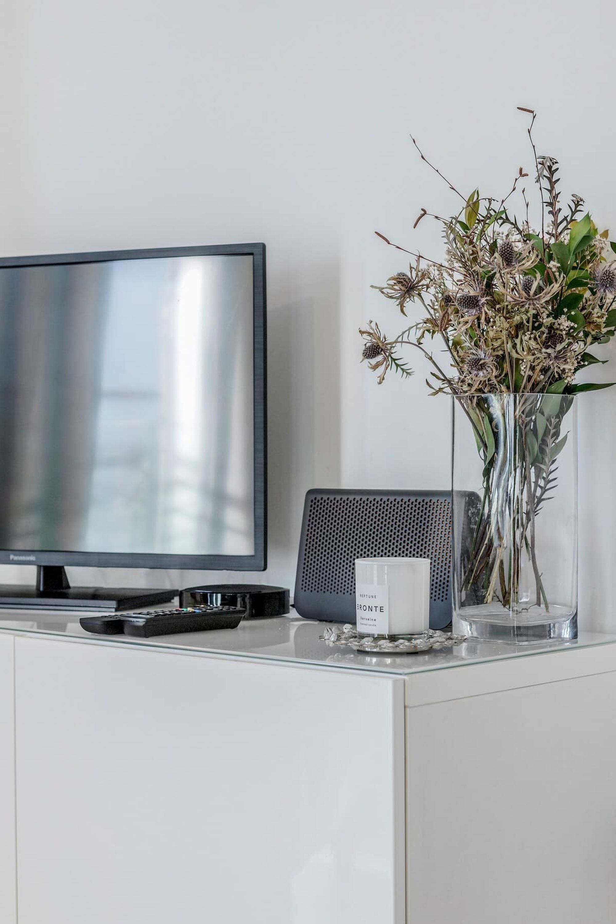 A flat-screen TV, a small speaker, and a vase with dried flowers on a white cabinet.