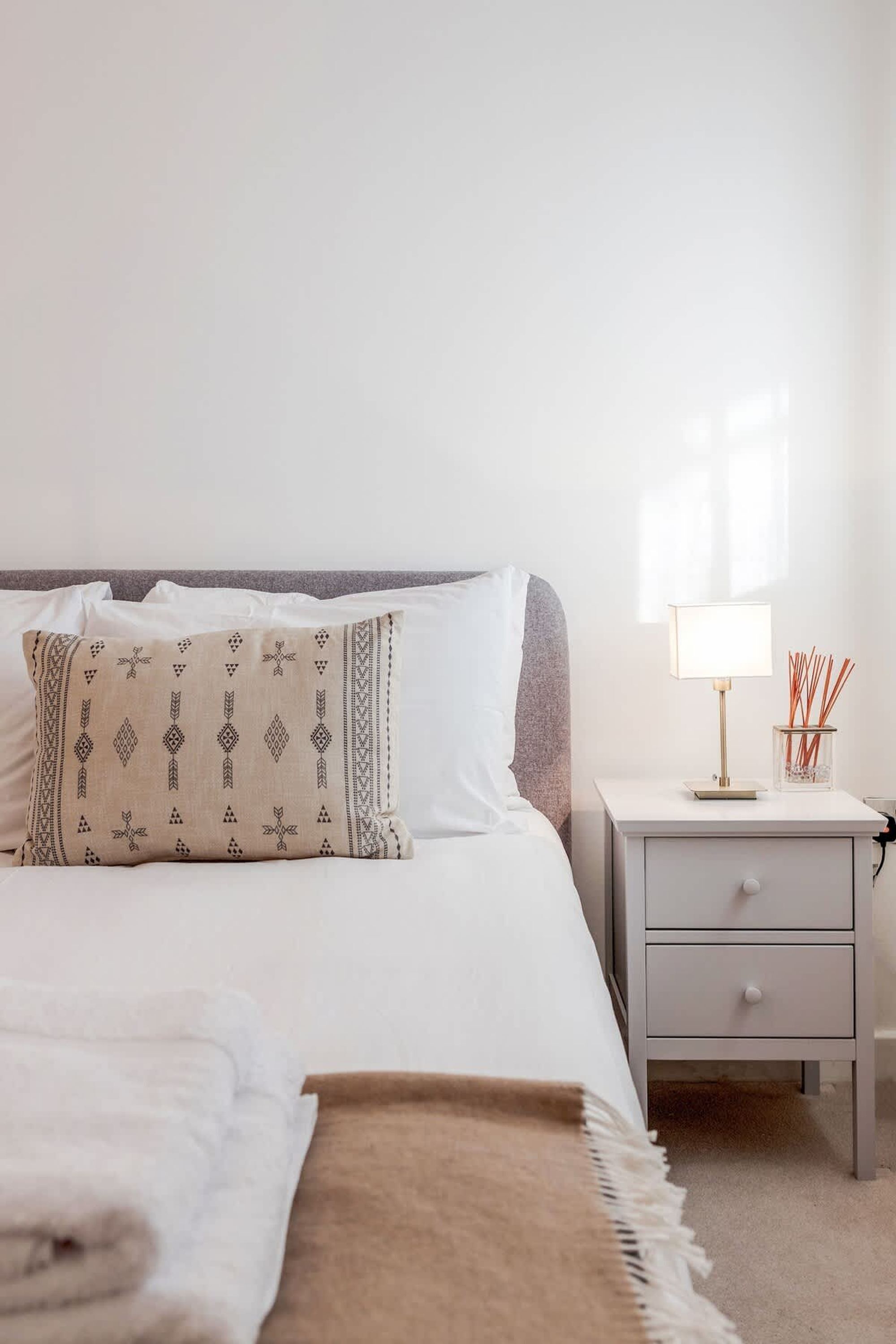 A neatly made bed with a patterned pillow, a bedside lamp, and a small container with reed sticks.