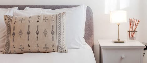 A neatly made bed with a patterned pillow, a bedside lamp, and a small container with reed sticks.