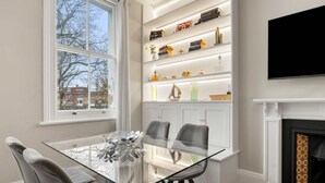 En moderne stue med et spisebord med glasplade, grå polstrede stole, en pejs og en reol med dekorative genstande.