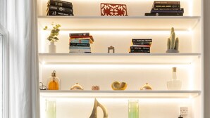 Et moderne interiør med et spisebord med glasplade, hvide reoler med bøger og pyngeting, og et minimalistisk design.