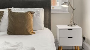 En pænt redt seng med et gråt sengegærde, hvide og brune puder samt et gråt tæppe. Et hvidt natbord med en lampe og et foldet hvidt håndklæde står ved sengen. Et vindue med udsigt til en murstensbygning er synligt.