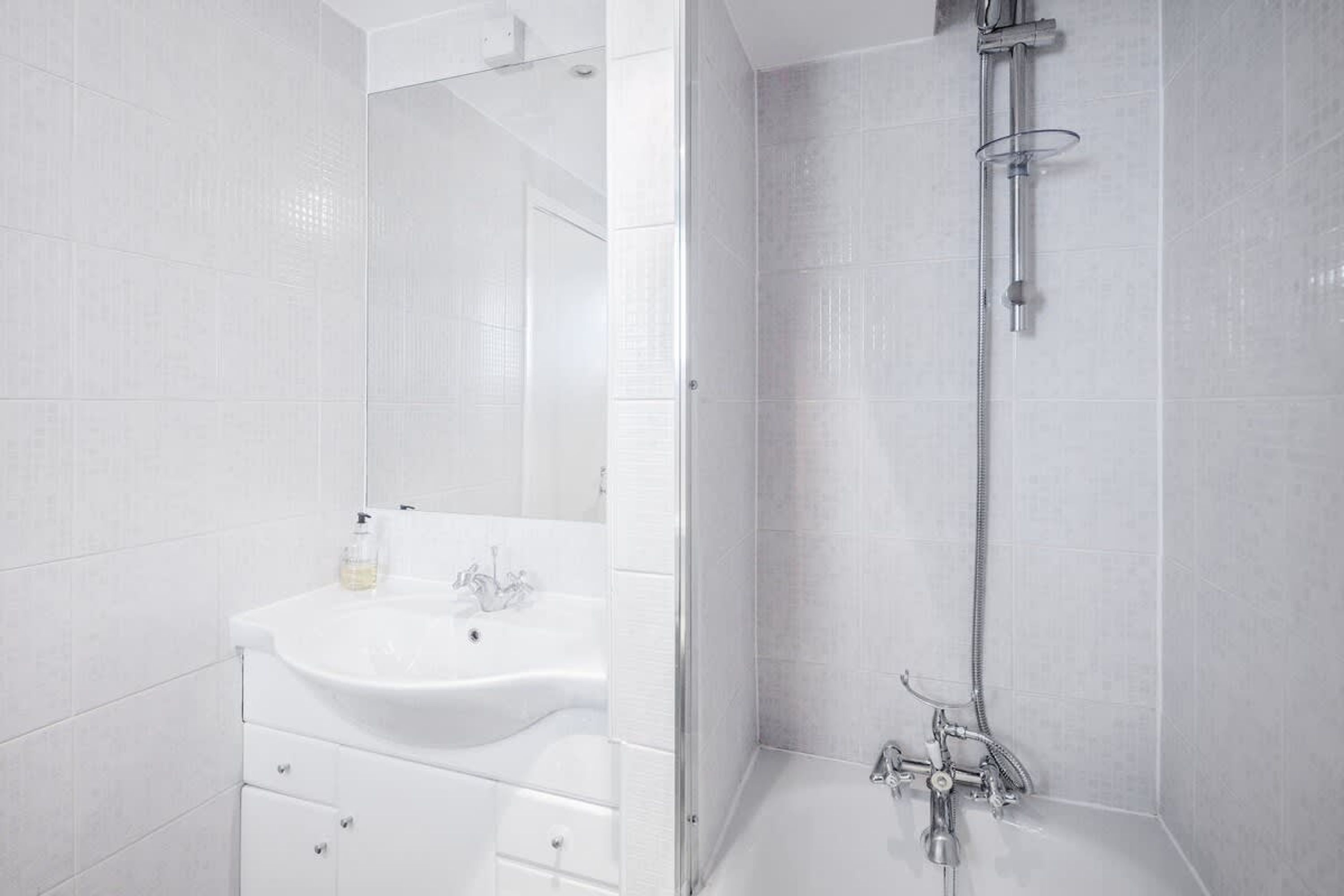 A bathroom with white tiles, a sink, a mirror, and a shower.