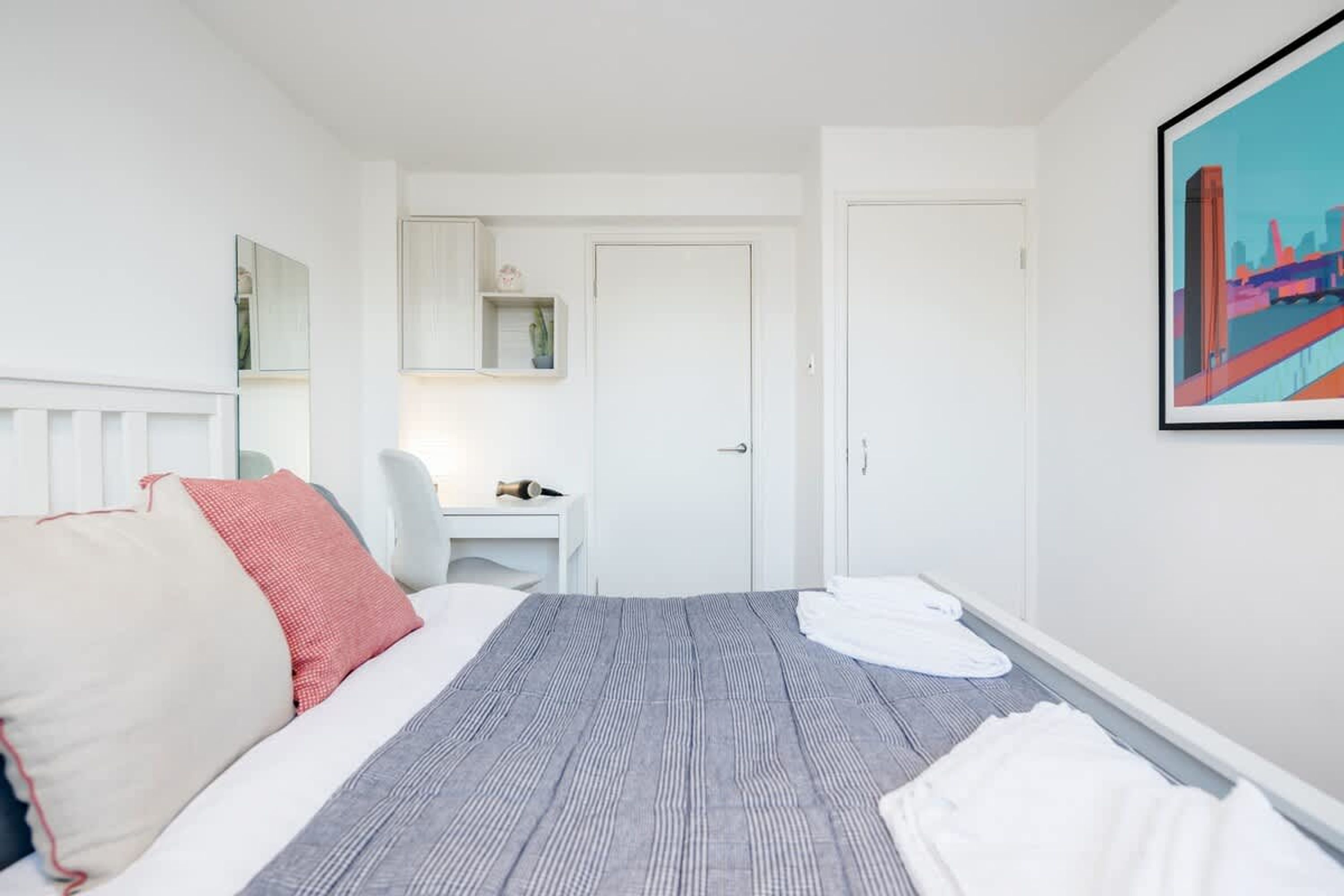 A hotel room with a bed, a desk, and a chair. There is a door and a painting on the wall.
