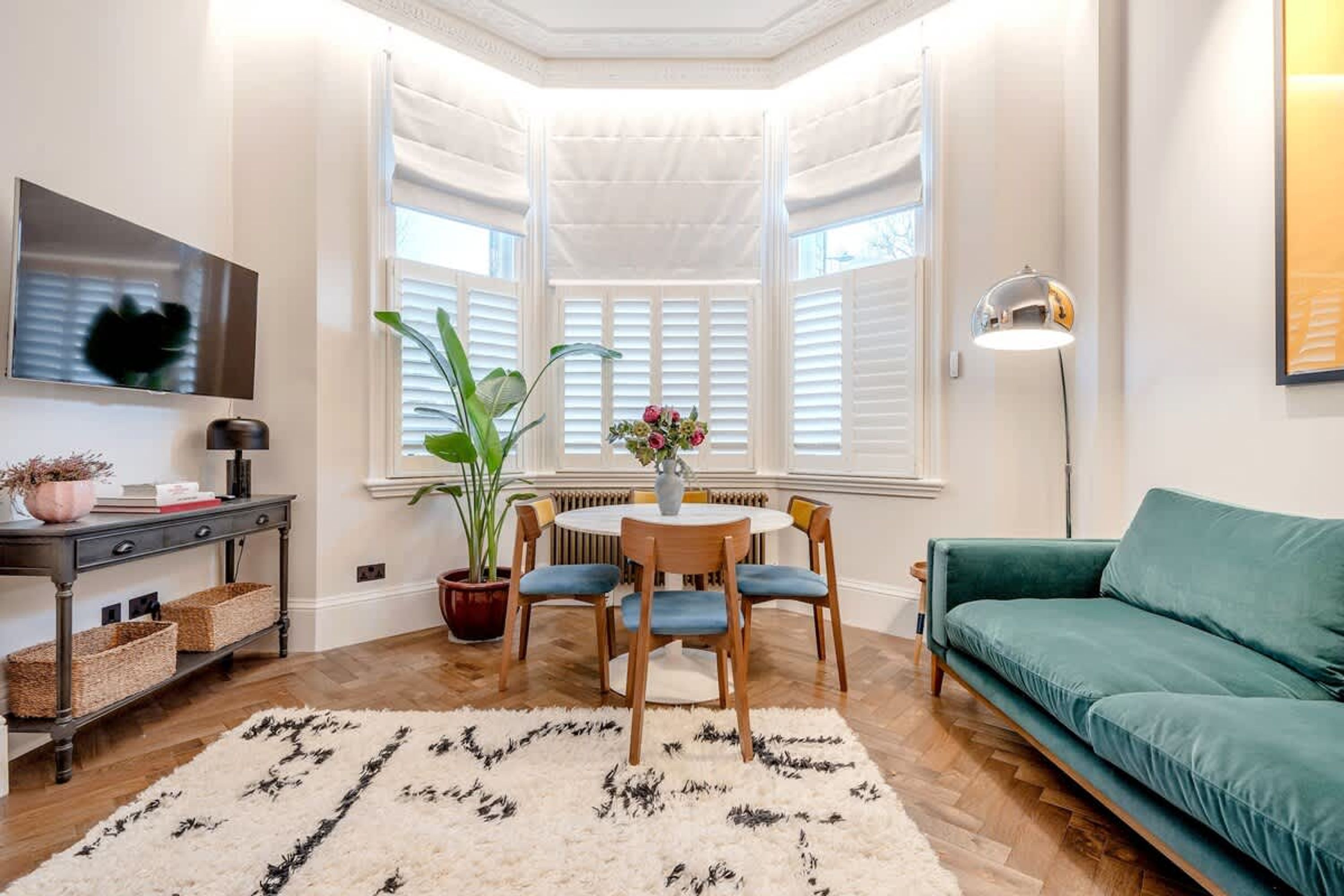 A cozy corner with a bay window, a dining table with chairs, a green sofa, a TV on the wall, and a potted plant.