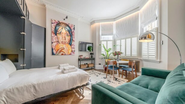 Une chambre à coucher moderne avec un lit, un canapé vert, une table à manger avec des chaises, une grande fenêtre avec des volets et un tableau accroché au mur.