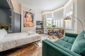 A modern bedroom with a bed, a green sofa, a dining table with chairs, a large window with shutters, and a painting on the wall. - Modern   spacious studio in Chelsea by King s Road (Greater London)