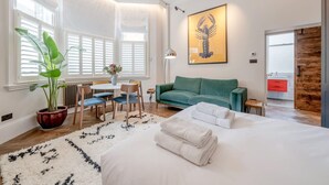 Une chambre d’hôtel moderne dotée d’un grand lit, d’un canapé vert, d’un coin repas avec une table et des chaises, et d’une grande fenêtre avec des stores.