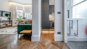 Une cuisine moderne avec des meubles sombres et un îlot central, un coin repas avec une chaise recouverte d’un tissu vert, et un porte-serviettes visible dans la salle de bain attenante.