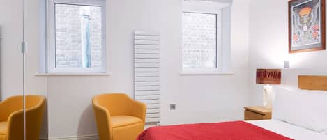 A bedroom with a bed, two chairs, a radiator, a nightstand, and a wall-mounted lamp.