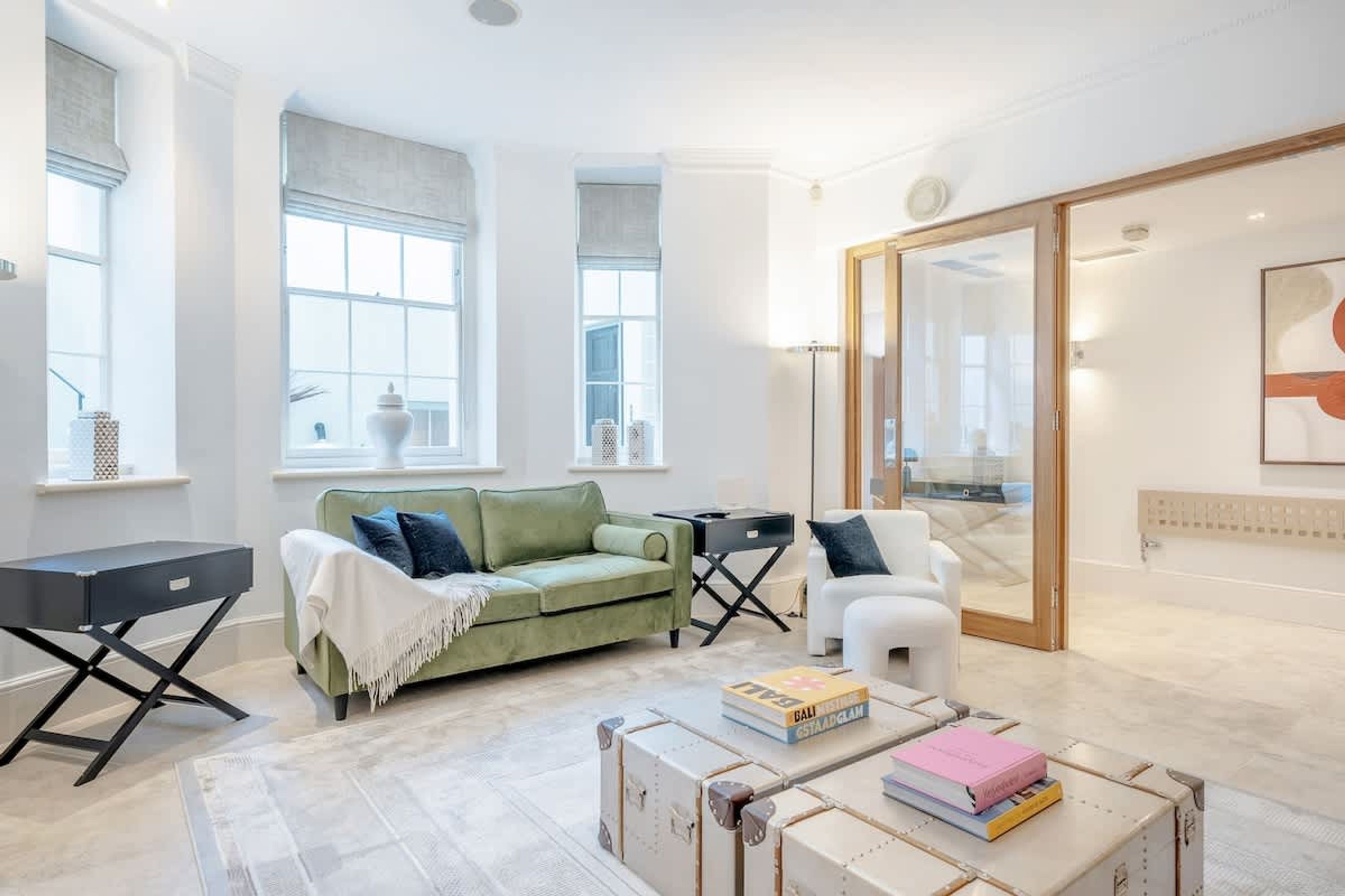 A modern living room with a green sofa, white armchair, and a coffee table with books. There are large windows and a sliding glass door leading to a balcony.