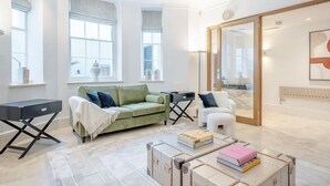 Ein modernes Wohnzimmer mit einem grünen Sofa, einem weißen Sessel und einem Couchtisch mit Büchern. Große Fenster und eine Glastür führen auf einen Balkon.