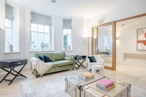 Ein modernes Wohnzimmer mit einem grünen Sofa, einem weißen Sessel und einem Couchtisch mit Büchern. Große Fenster und eine Glastür führen auf einen Balkon.