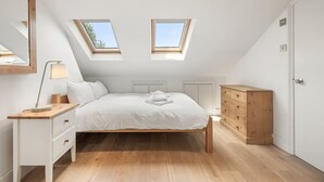 Ein Schlafzimmer mit schrägen Wänden, Dachfenstern, einem Bett, einem Nachttisch mit Lampe, einer Kommode und einer Tür.