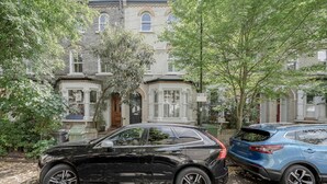 Ein schwarzer SUV parkt vor einem historischen Gebäude mit einem Erkerfenster, neben ihm steht ein blauer SUV.