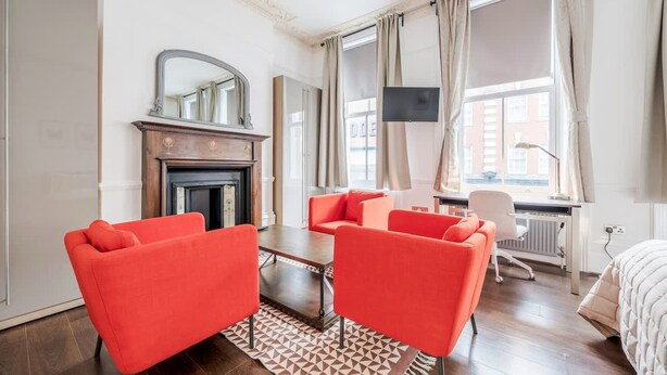 A modern hotel room with a fireplace, two red armchairs, a coffee table, a TV, and a large window with curtains. - Unbeatable location in Camden-spacious studio flat (Greater London)