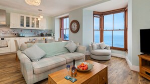 Un salón con sofá, una mesa de centro con frutas y vasos, un mueble para televisión y vistas al mar a través de amplias ventanas.