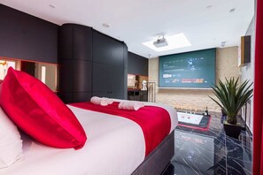 A modern hotel room with a large bed, a flat-screen TV, a ceiling projector, and a small balcony with a plant. - Adresse Secrete Suite Spa Prestige avec Balneo (Dijon)