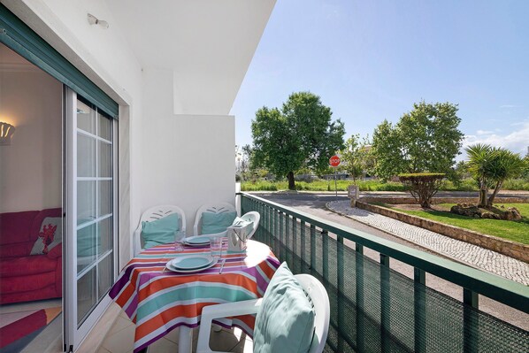 A balcony with a table set for dining, a red sofa, and a view of the street and greenery. - Holiday Home 'Zuri' with Balcony and Wi-Fi (União das freguesias de Conceição e Cabanas de Tavira)
