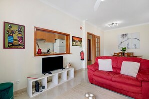 Un salon avec un canapé rouge, un téléviseur posé sur un meuble blanc, un coin cuisine avec un réfrigérateur et une table à manger avec des chaises.