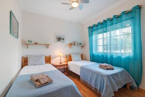 Une chambre avec deux lits, un ventilateur de plafond, une fenêtre avec des rideaux bleus et des étagères murales ornées de tableaux.