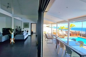 A modern living room with a sofa, a coffee table, and a TV. There is a balcony with a pool and a view of the ocean. - Villa Rêve Austral - Three Bedroom Villa, Sleeps 6 (Saint Pierre)