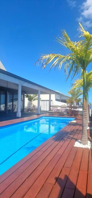 A swimming pool with wooden decking, a palm tree, and a building with large windows. - Villa Rêve Austral - Three Bedroom Villa, Sleeps 6 (Saint Pierre)