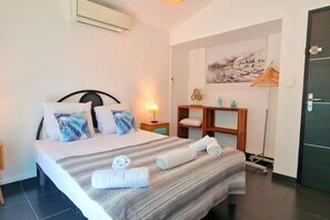 A neatly made bed with pillows, a bedside lamp, a small wooden shelf with towels, and a painting on the wall. - Villa Rêve Austral - Three Bedroom Villa, Sleeps 6 (Saint Pierre)
