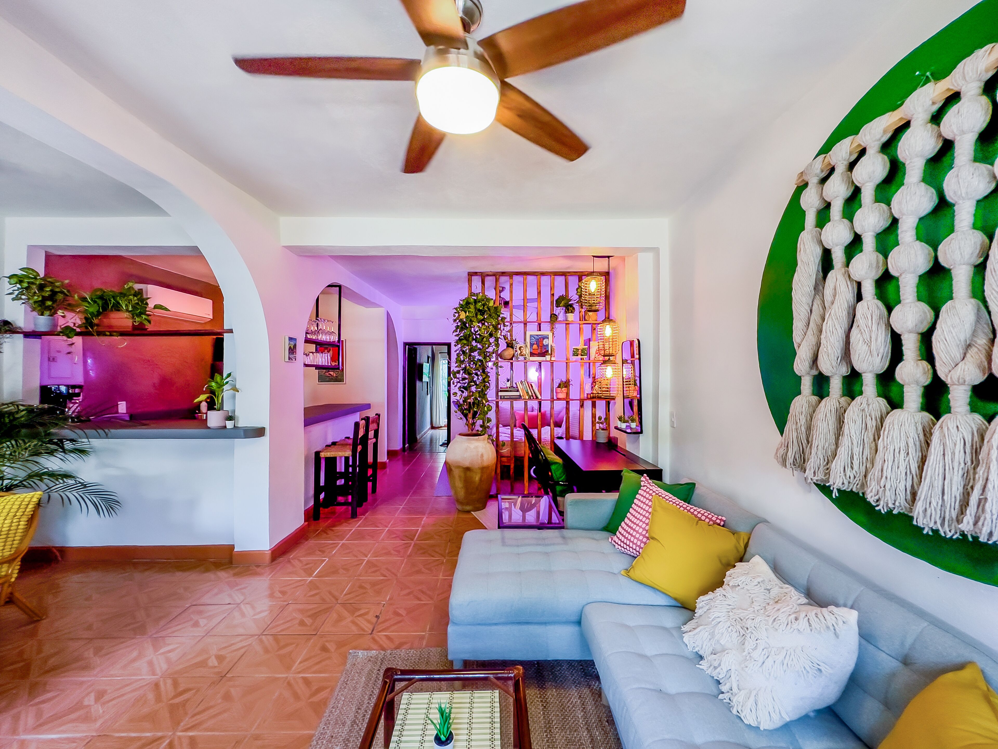 A living room with a blue sofa, a coffee table, and a ceiling fan. There is a kitchen area with a pink wall and a dining area with chairs and a table.