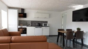 A modern living room with a brown leather sofa, a dining area with a wooden table and chairs, a kitchen with white cabinets and a black TV mounted on the wall. - 4 Star Holiday Home in Aabybro (Aabybro)