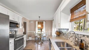 Une cuisine moderne avec des meubles blancs, des appareils en acier inoxydable, un coin repas avec une table et des chaises, et une fenêtre donnant sur un paysage verdoyant.