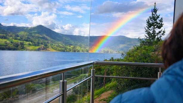 Une personne est assise près d’une fenêtre donnant sur un lac, avec un arc-en-ciel, des montagnes et un ciel dégagé.