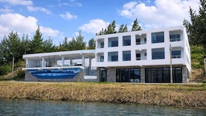 Une maison moderne à deux étages, dotée d’un grand balcon donnant sur une étendue d’eau et entourée d’arbres.