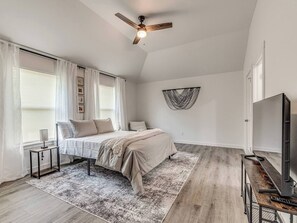 A bedroom with a bed, a ceiling fan, a wall-mounted TV, and a large window with curtains.