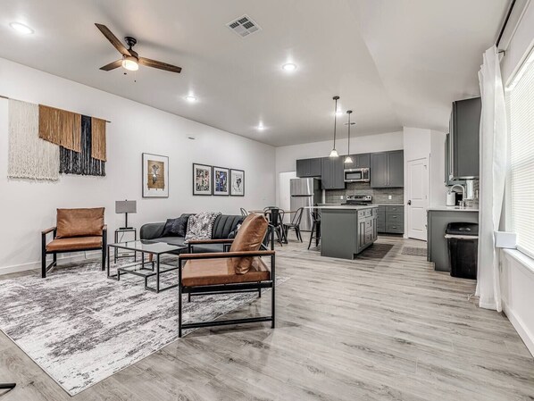 A spacious living area with a sofa, chairs, a coffee table, and a rug. A kitchen with modern appliances is visible in the background.