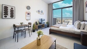 A modern living room with a dining area, a large window with a city view, and a bed with a view. - Stylish Studio with Balcony & Pool View in Arjan – Close to Miracle Garden (Dubai)