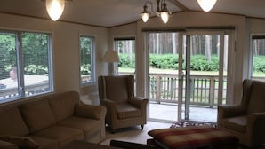 A living room with a sofa, two armchairs, a coffee table, and a lamp. There are large windows and a sliding glass door leading to a deck. - Living The Dream (Holt)