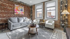 A living room with a brick wall, a gray sofa, white armchairs, a round coffee table, and a view of buildings through large windows. - Park Hotel 2BR Suite 406 (Chattanooga)